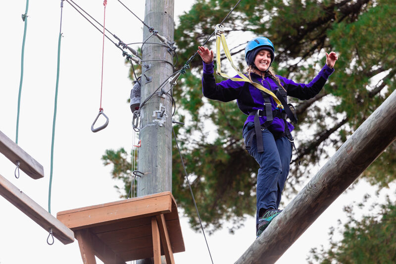A woman wearing a blue helmet and a harness is carefully walking on a wooden beam, part of a high ropes course. She is holding her arms out for balance and appears focused on her next step. The course is set among trees, with a gray pole and various ropes and platforms visible in the background. The sky is overcast, providing soft lighting for the scene.